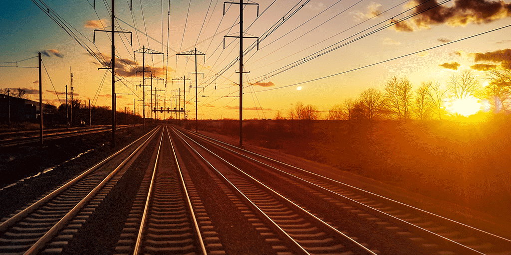 Train tracks leading towards the horizon with overhead wires and a sunset.